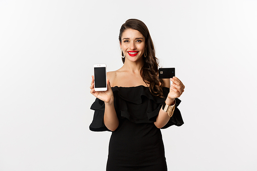 Online shopping concept. Fashionable brunette woman in black dress, showing mobile screen and credit card, smiling pleased, standing over white background.