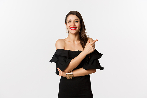 Fashion and beauty. Elegant woman with red lips, black dress, smiling and pointing finger right at logo, standing over white background.