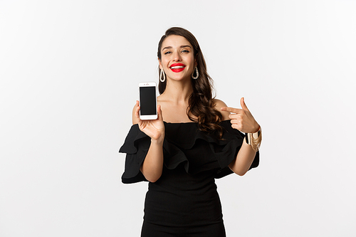 Online shopping concept. Fashionable woman in black dress pointing finger at smartphone screen, showing application, standing over white background.