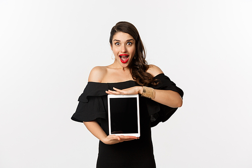 Fashion and shopping concept. Amazed young woman showing online website promo offer on tablet screen, looking excited camera, standing in black dress, white background.