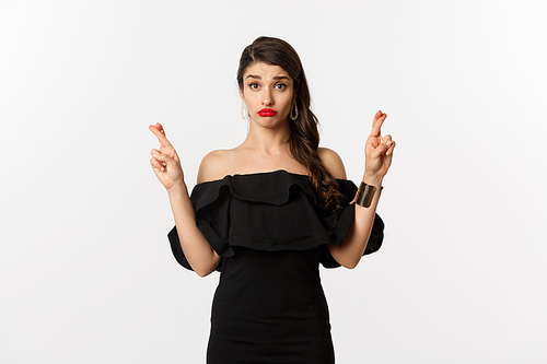 Fashion and beauty. Hopeful silly woman in black dress making wish, holding fingers crossed for good luck, standing over white background.