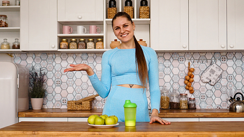 Athletic woman talks about healthy eating in the kitchen and laughs. On the table are green apples and a green bottle for a sports drink and water