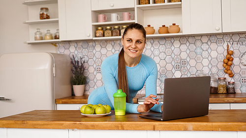Smiling woman using computer in modern kitchen interior. Cooking and healthy lifestyle concept. A woman is looking at the camera and smiling