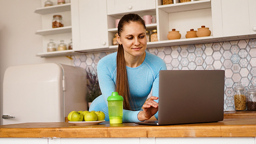 Smiling woman using computer in modern kitchen interior. Cooking and healthy lifestyle concept. A woman is looking for a recipe or is streaming online