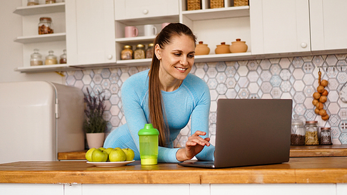 Smiling woman using computer in modern kitchen interior. Cooking and healthy lifestyle concept. A woman is looking for a recipe or is streaming online