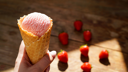 Close up image of woman hand holding fresh waffle cone with raspberry ice cream on the wooden background. Berries on a blurred background.