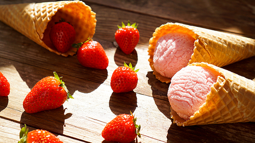Strawberry ice cream in a waffle cone. Red berries and ice cream balls on a wooden background. Bright light. Shade from the sun. Summer photo.