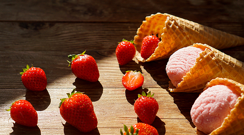 Strawberry ice cream in a waffle cone. Red berries and ice cream balls on a wooden background. Bright light. Shade from the sun. Summer photo.