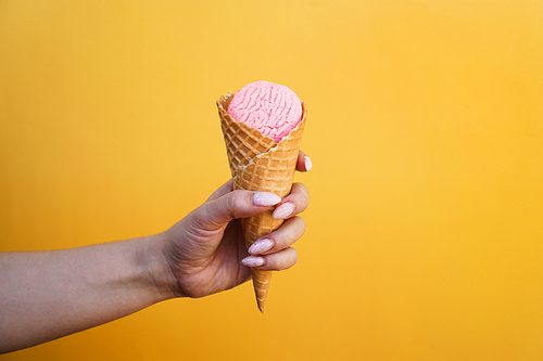 Ice cream in waffle cup in female hand on yellow background. Pink strawberry ice cream in a waffle cone