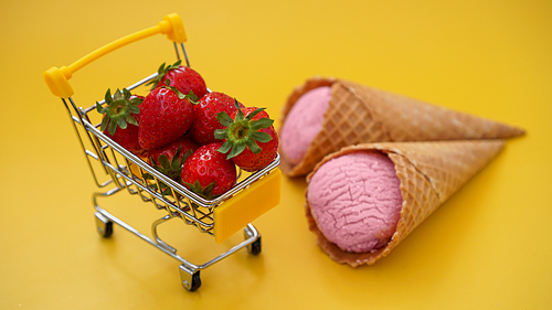 Fresh strawberries in a shopping cart and strawberry ice cream on a bright yellow background. Buying berries for making ice cream