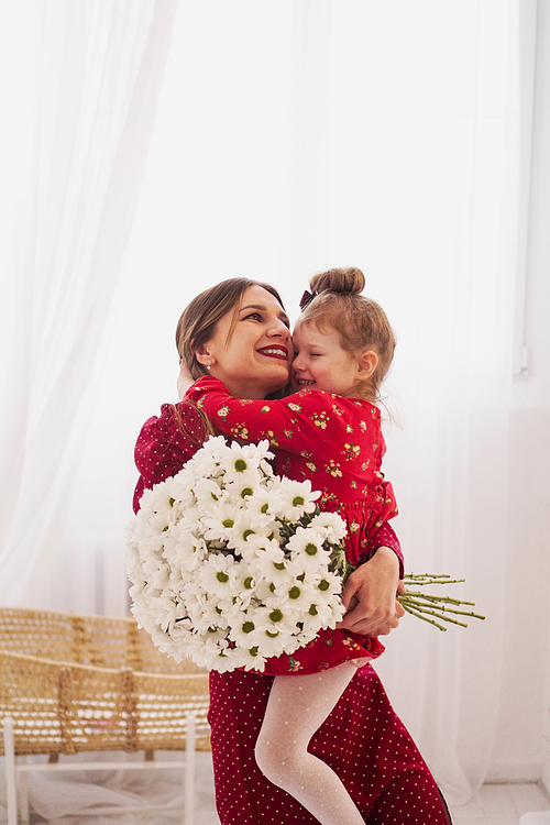 Mom and little daughter in red dresses in a bright bedroom in a Scandinavian style. Happy family. Mom holds a bouquet of daisies - spring photo