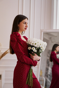 Beautiful brunette in a red dress with a bouquet of daisies at home. Spring photo.