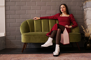 Beautiful brunette woman in a red dress and white shoes on a green sofa. Gray brick wall in the interior