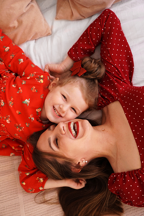 Mom and her little daughter lie on the bed facing each other. They are happy. Both are wearing red dresses.