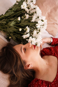 A beautiful brunette in a red dress lies on the bed next to a bouquet of daisies. Portrait of a beautiful woman.