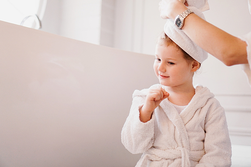 Mom puts a towel on her daughter's hair. Little daughter in a white bathrobe in a bright bathroom.