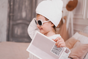 Smiling little girl in a white bathrobe after a bath. White cozy interior. Hygiene and baby fashion concept. She is wearing sunglasses, the girl is holding a magazine in her hand
