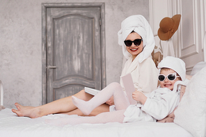 Mother and daughter in the bedroom in bathrobes. They read magazines lying on the bed. They are in sunglasses looking at the camera and laughing. Focus on mom's face