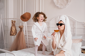 Mother and daughter in the bedroom in bathrobes. Happy daughter is jumping on the bed. Mom is drinking a glass of white wine. Funny motherhood fatigue concept