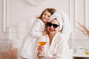 Mother and daughter in the bedroom in bathrobes. The girl hugs her mom. Mom is holding a glass of white wine.