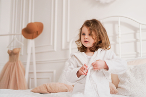 Smiling little girl in a white bathrobe after a bath. White cozy interior. Hygiene and baby fashion concept