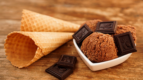 Homemade chocolate ice cream with chocolate pieces in a white bowl. Wooden background and two waffle cones
