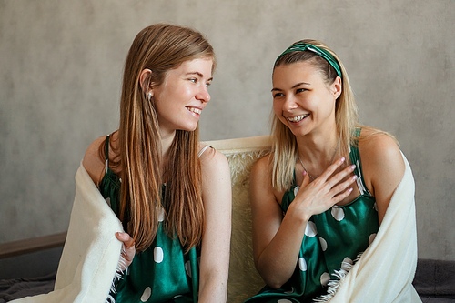 Two laughing young woman having fun when hiding under white blanket on bed in bedroom. Pretty girlfriends during pajamas or sleepover party