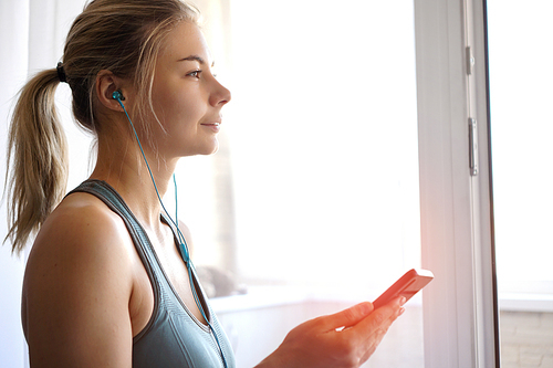 Happy young pretty sporty girl stands near the window and listens to music on headphones from smartphone