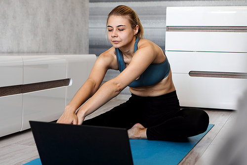 Pretty young woman in sportswear watching online video on laptop and doing exercises at home. Distant training with personal trainer, online education