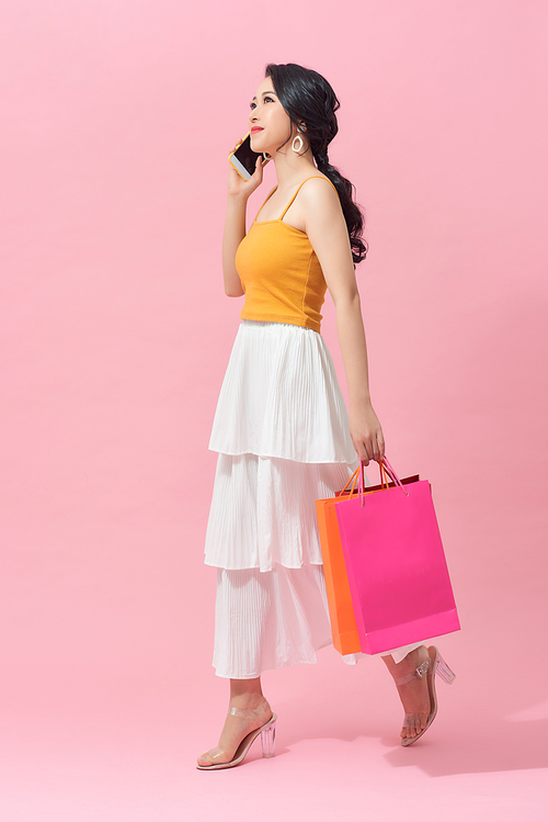 Full length of a beautiful young woman wearing colorful clothes standing isolated over pink background, carrying shopping bags, holding mobile phone