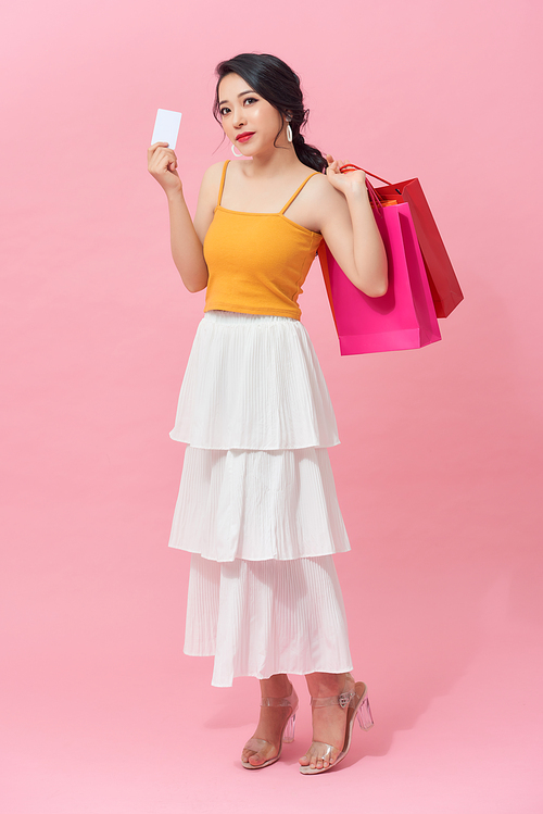 Full length of beautiful young Asian with colorful shopping bag and credit card over pink background.
