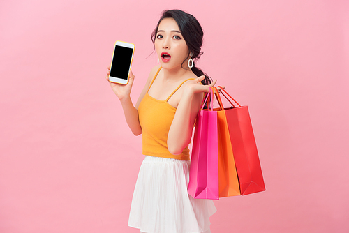 Photo of shocked woman 20s in dress looking at smartphone in hand with surprise while holding shopping bags isolated over pink background