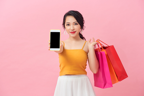 Portrait of a pretty excited girl with colorful shopping bags showing blank screen mobile phone over pink background.