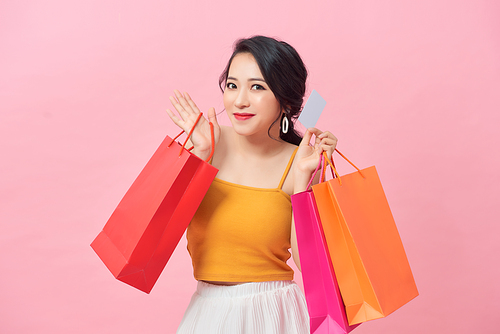 Close-up portrait of happy young Asian woman holding credit card and colorful shopping bags, isolated on pink background