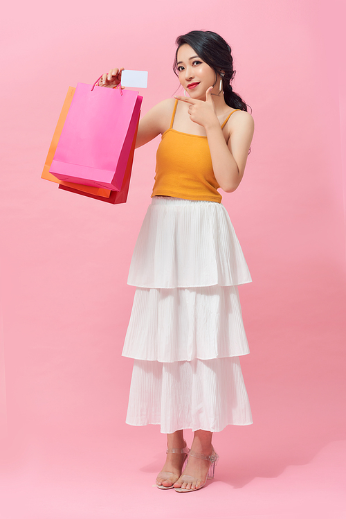 Full length of excited Asian woman with colorful shopping bag while standing over pink background.