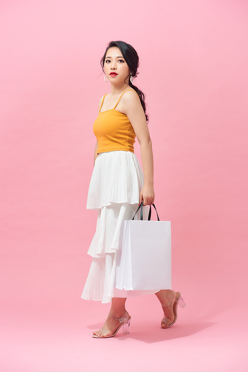 Full length of young woman holding shopping bags and smiling while walking isolated over pink background.