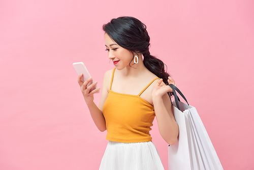 Beautiful girl is holding shopping bags, using a smart phone over pink background.