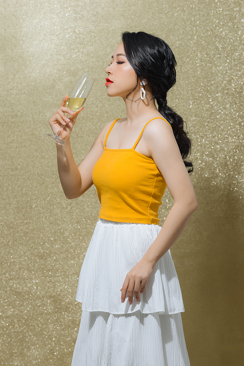 Portrait of cheerful young Asian woman drinking wine isolated over gold background.