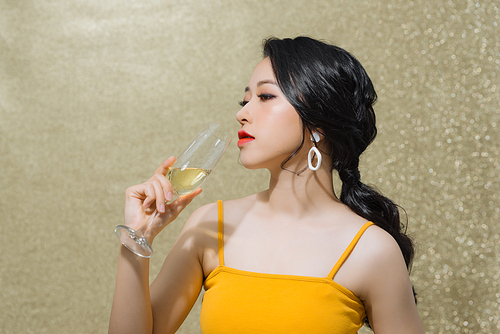 Portrait of cheerful young Asian woman drinking wine isolated over gold background.