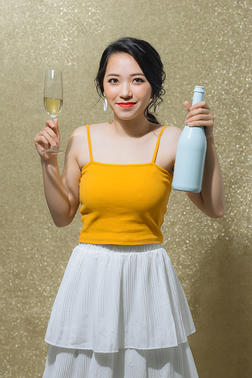 Portrait of happy young Asian woman holding champagne bottle and glass over gold background. Party concept.