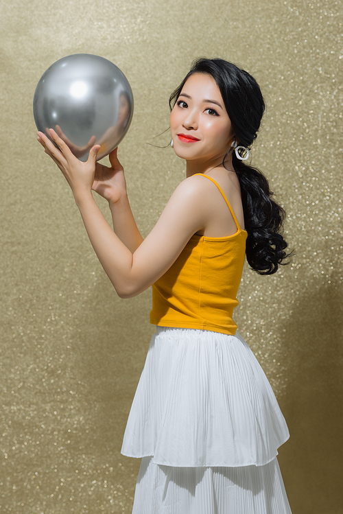 Beautiful young Asian woman holding ballon when standing isolated over gold background.