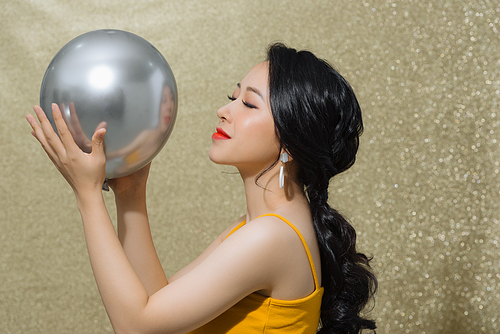 Cheerful young Asian woman with balloons laughing over gold background.