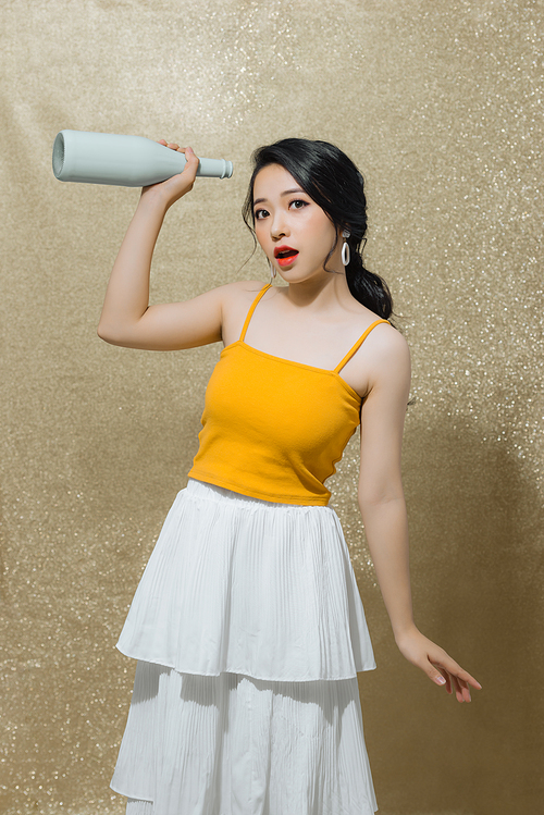 Portrait of sweet, gorgeous young Asian woman with bottle of beverage in hands isolated on bright gold background