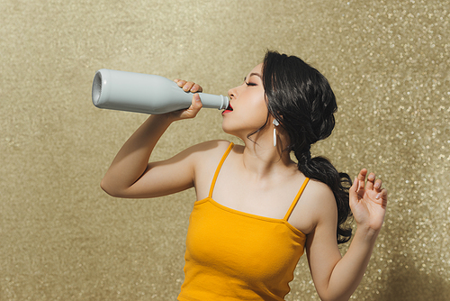 Attractive young Asian woman drinking wine wit bottle over gold background.