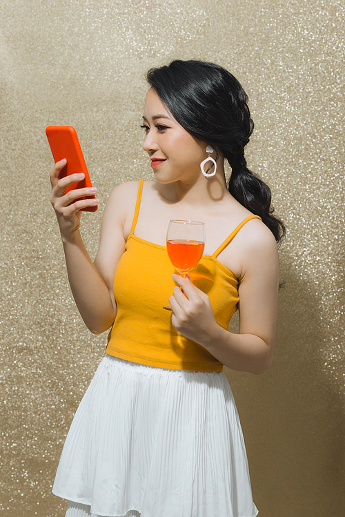 Attractive young Asian woman using phone and holding wine glass isolated over gold background.