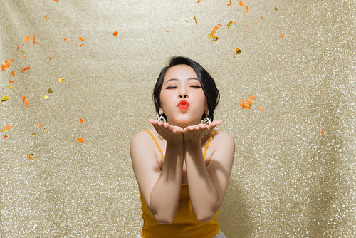 Portrait of beautiful young woman with party whistle and falling confetti on color background