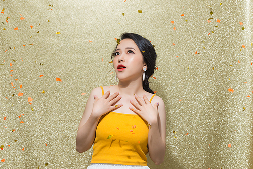 Portrait of a cheerful beautiful girl wearing dress standing under confetti rain and celebrating isolated over gold background