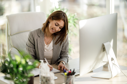 A smiling business woman write something down in a notebook while using a computer in her home office.