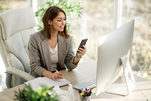 An attractive business woman using smartphone while working at computer in her home office.