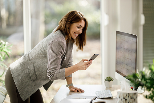 A smiling business woman using smartphone while working at computer in her home office.